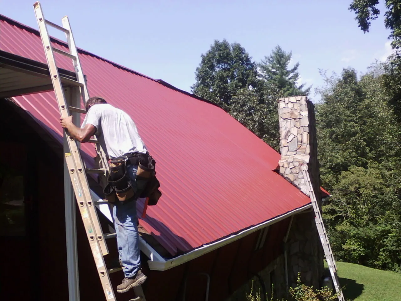 Roofer on a ladder beside a red metal roof and stone chimney — Soffit & Fascia Repair by Roofing Brothers in Clayton
